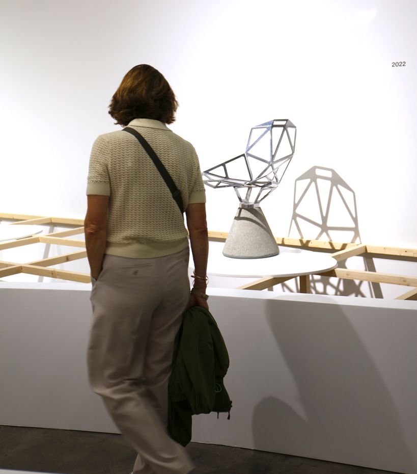 Une femme de dos, elle regarde une chaise posée sur un socle blanc. L'ombre de la chaise au design contemporain et formée de découpes donne une effet de réplique sur le mur