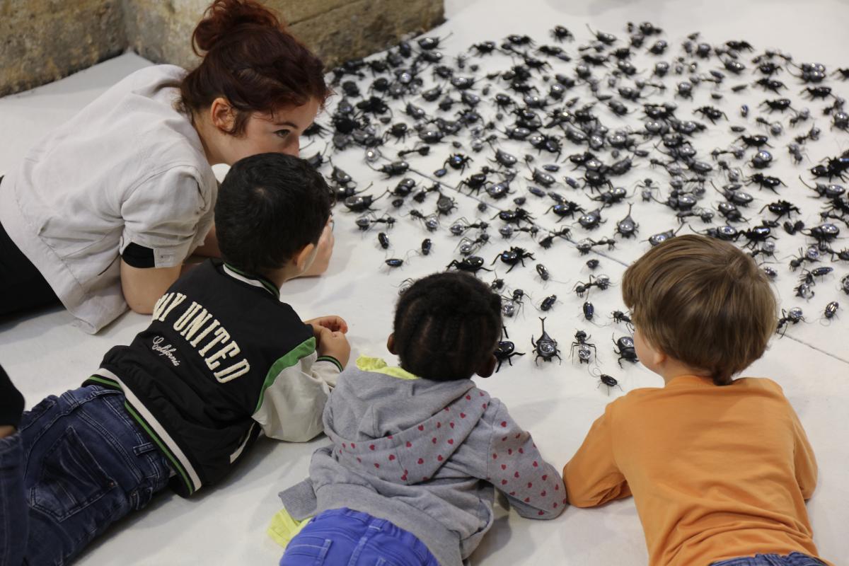 Des enfants et une médiatrice allongés au sol sur le ventre observent une œuvre