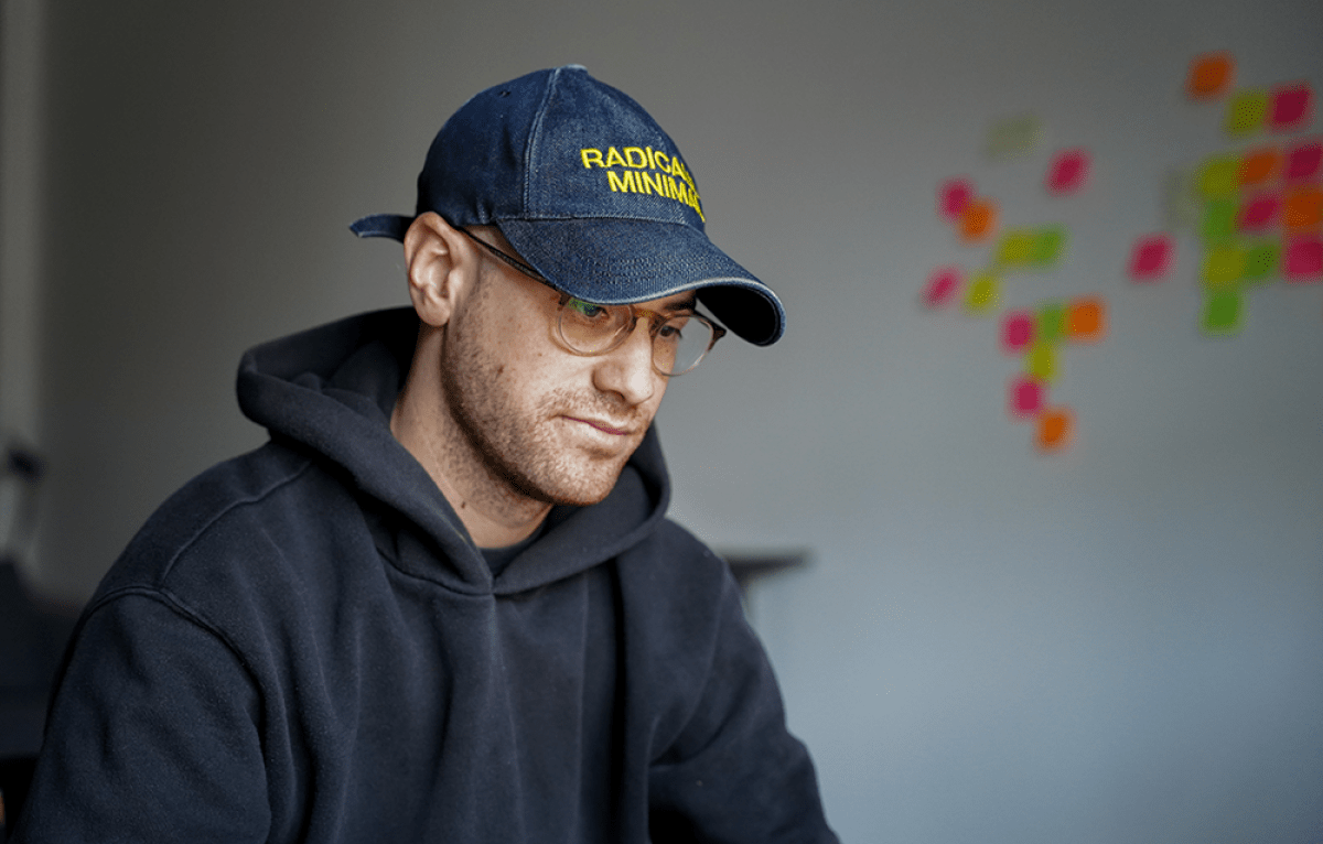 Portrait un jeune homme vêtu d'un sweet-shirt à capuche gris et d'une casquette bleu foncée. En arrière plan sur un mur blanc on distingue une constellation de post-it de couleur 